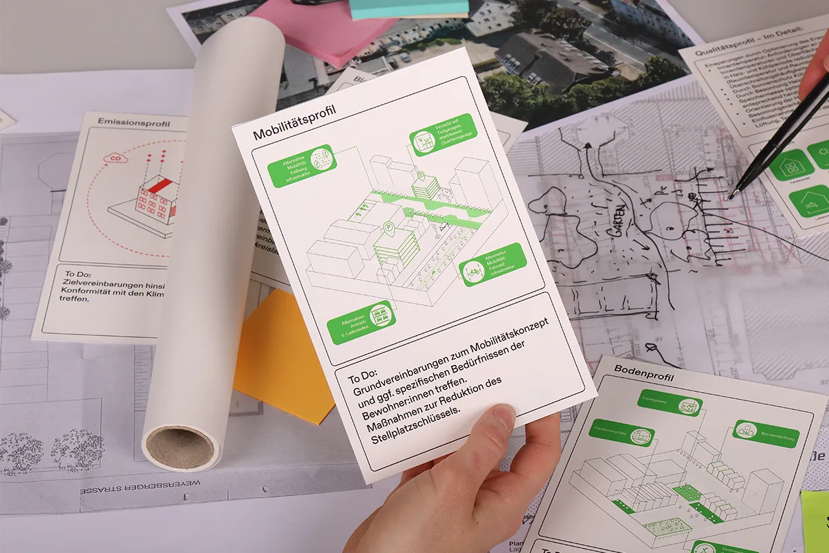 One workshop participant holds the action card Mobilitaetsprofil while another points to a urban plan sketch